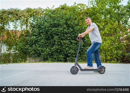 Senior man riding driving kick push scooter on the asphalt in front of the bushes green fence in a summer day