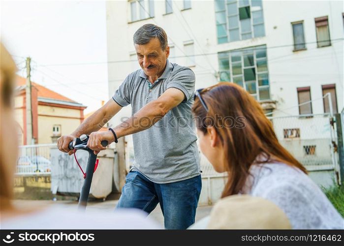 Senior man riding electric kick scooter while talking to some woman in neighborhood by the street in summer day