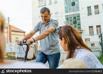 Senior man riding electric kick scooter while talking to some woman in neighborhood by the street in summer day