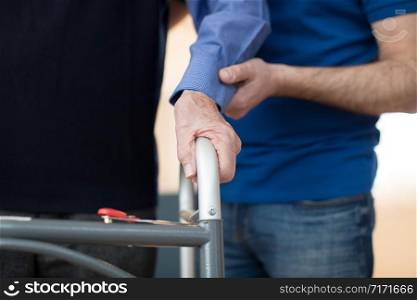 Senior Man&rsquo;s Hands On Walking Frame With Care Worker In Background