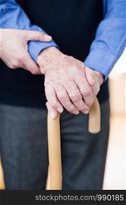 Senior Man's Hands On Walking Stick With Care Worker In Background