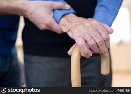Senior Man's Hands On Walking Stick With Care Worker In Background