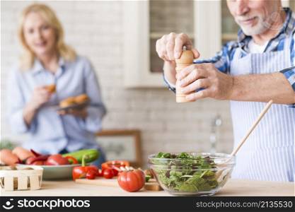 senior man seasoning green vegetables salad her wife holding muffins hand backdrop