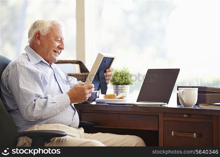 Senior Man Sitting At Desk Looking At Photo Frame
