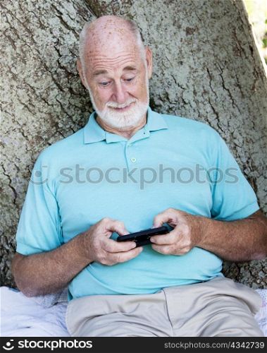 Senior man sitting outdoors under a tree, sending a text on his smart phone.