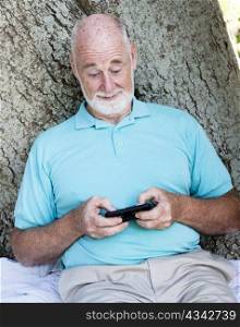 Senior man sitting outdoors under a tree, sending a text on his smart phone.