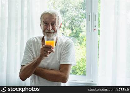 senior man smile and hold glass of orange juice stand, in front of window in white room