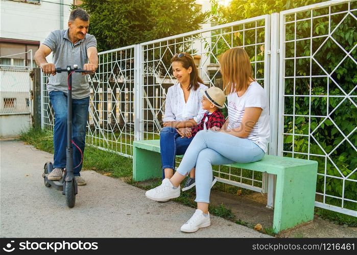 Senior man standing on the electric scooter by the bench with his family mother neighbor sitting on the bench by the fence in summer day