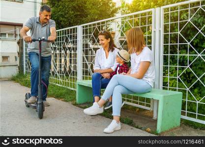 Senior man standing on the electric scooter by the bench with his family mother neighbor sitting on the bench by the fence in summer day
