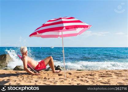 Senior man sun bathing at the beach under parasol with waves, sea and sand