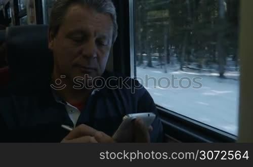 Senior man traveling by train and typing text message on smartphone using pen. From time to time he looking at winter scenery through the window