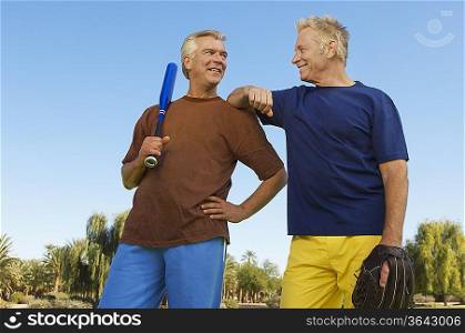 Senior man with baseball mitt leaning on man with baseball bat, outdoors