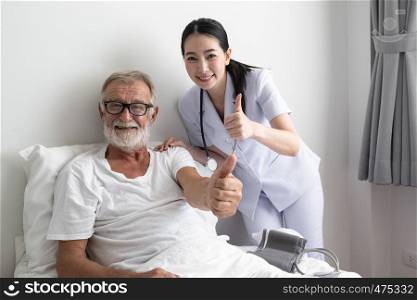senior man with smiling nurse, takes care and discussion after health check by thumb up cheer on bed at nursing home