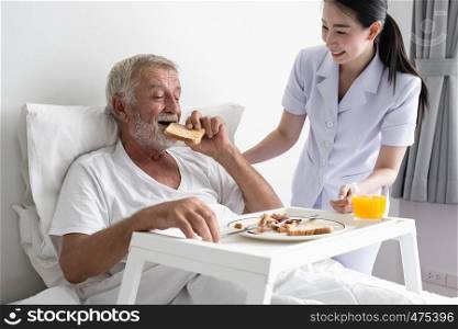 senior man with smiling nurse, takes care breakfast and discussion and cheer on bed at nursing home