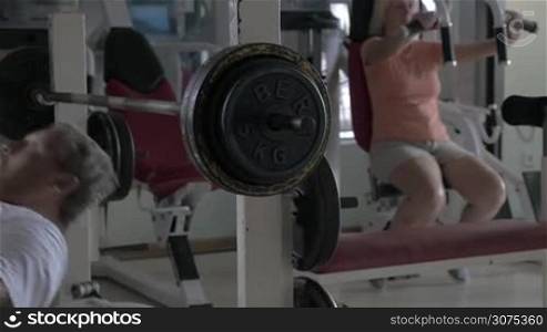 Senior man working out with bar-bell lifting, woman training on exerciser in background