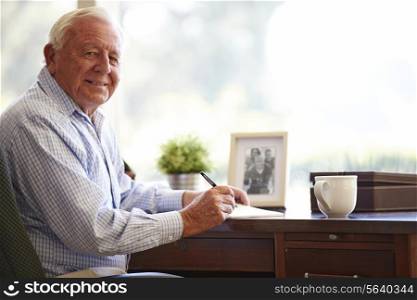 Senior Man Writing Memoirs In Book Sitting At Desk