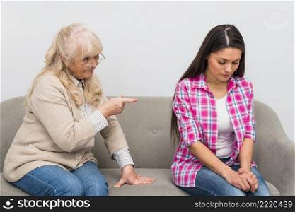 senior mother scolding her adult daughter sitting sofa against wall
