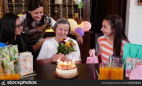 Senior mother with daughter and granddaughters with flowers and present at birthday party. Celebrating life and love concept between generations. Mothers day concept. Slow motion hand held movement