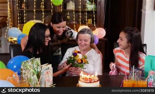 Senior mother with daughter and granddaughters with flowers and present at birthday party. Celebrating life and love concept between generations. Mothers day concept. Slow motion hand held movement