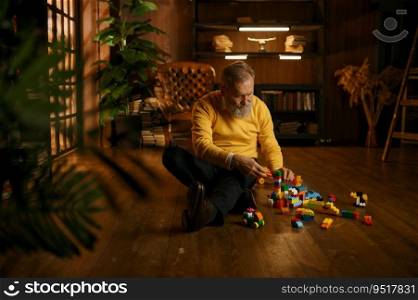 Senior old man playing toy building blocks while sitting on home floor during cozy evening. Handsome adult businessman on retirement. Senior old man playing toy building blocks while sitting on home floor