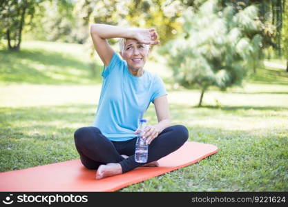 Senior sportive woman sitting on mat outside and resting after workout