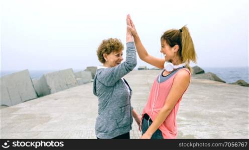 Senior sportswoman and female friend high five by sea pier. Senior sportswoman and female friend high five