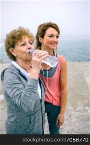 Senior sportswoman drinking water with female coach by sea pier. Senior sportswoman drinking water with female coach