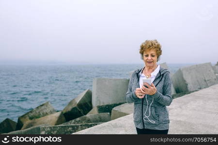 Senior sportswoman with headphones looking at her smartphone by sea pier. Senior sportswoman looking at her smartphone