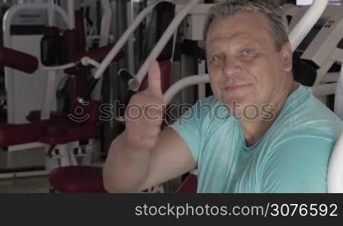 Senior strong man giving thumb up with smile in fitness centre against gym machine