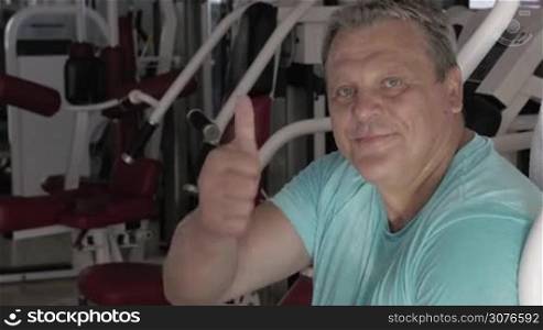Senior strong man giving thumb up with smile in fitness centre against gym machine