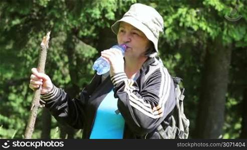Senior tourist woman walking and drinking water in the forest in summer.