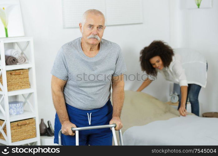 senior using a zimmer while carer in making the bed