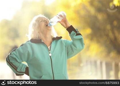 Senior Woman Drinking Water after Jogging Outdoors in the Bright Autumn Evening