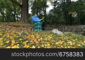 Senior woman enjoying his Retirement in chair under autumn tree, reading book Dolly shot, Wide Angle, Side View