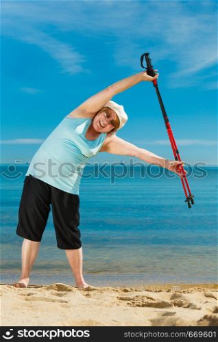 Senior woman enjoying nordic walking, doing warmup exercises with poles on sea shore, sunny summer day. Health, activity in old age.. Senior woman warming up with walking poles