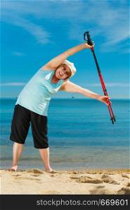 Senior woman enjoying nordic walking, doing warmup exercises with poles on sea shore, sunny summer day. Health, activity in old age.. Senior woman warming up with walking poles