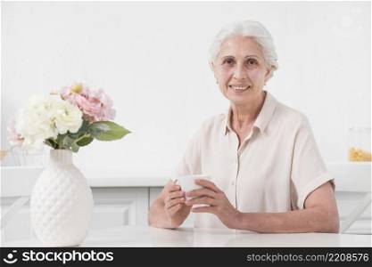 senior woman holding cup coffee with flower vase white table