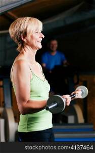 Senior woman in a gym exercising with barbell; in the background her friends