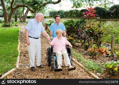 Senior woman in a wheelchair being walked through the nursing home garden by an orderly and her husband.