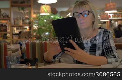 Senior woman in glasses spending evening in cafe and passing time with surfing internet on digital tablet