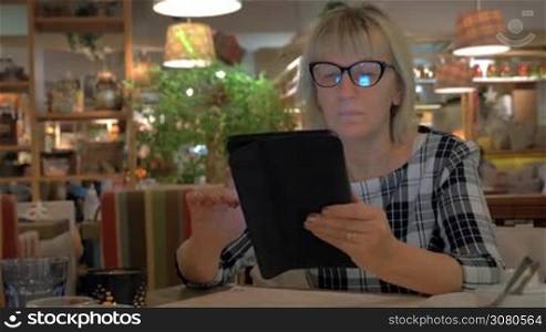 Senior woman in glasses spending leisure time in cafe and browsing web on digital tablet