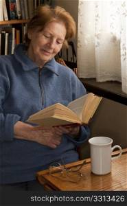 senior woman in her eighties reading a book by window in a study or library