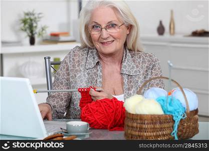 Senior woman knitting in front of her laptop computer