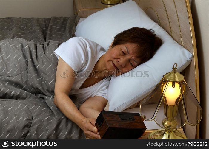 Senior woman looking at alarm clock during night time. Insomnia concept.