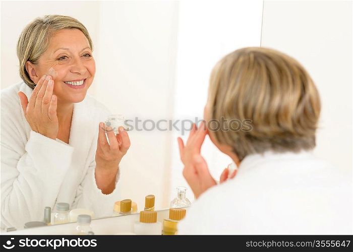 Senior woman looking in bathroom mirror and applying anti-wrinkles cream