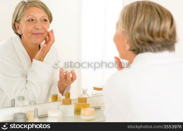 Senior woman looking in bathroom mirror applying anti-wrinkles face cream