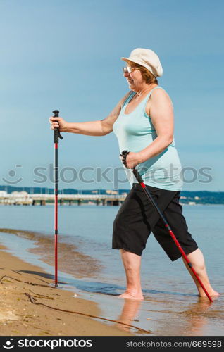 Senior woman practicing nordic walking on sea shore, Active elderly female enjoying sunny summer day. Healthy lifestyle in old age.. Senior woman practicing nordic walking on beach