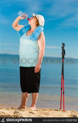 Senior woman practicing nordic walking on sea shore, drinking fresh water after activity. Elderly female enjoying sunny summer day. Healthy active retirement age.. Senior woman practicing nordic walking on beach