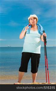Senior woman practicing nordic walking on sea shore, drinking fresh water after activity. Elderly female enjoying sunny summer day. Healthy active retirement age.. Senior woman practicing nordic walking on beach