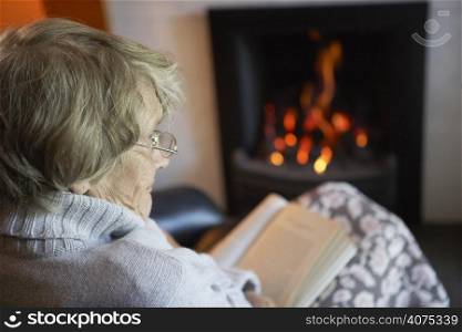 Senior Woman Reading Book By Fire At Home